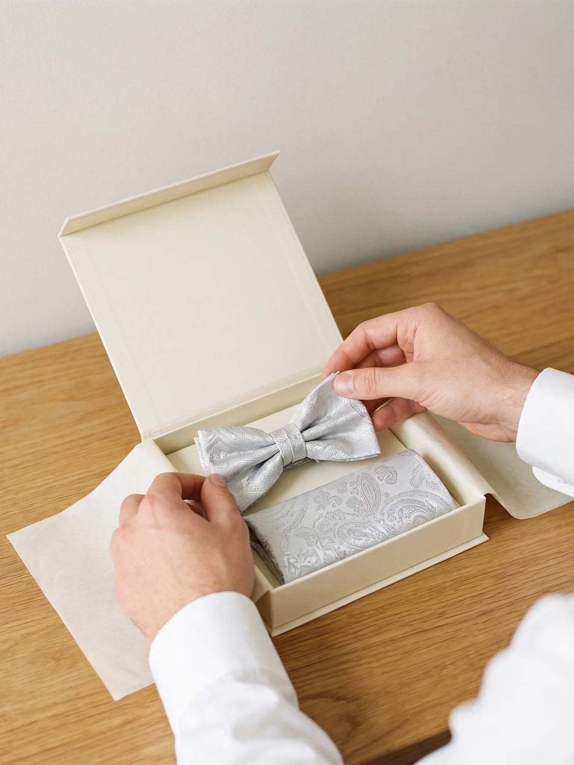 Hände öffnen eine cremefarbene Geschenkbox mit Fliege und Einstecktuch